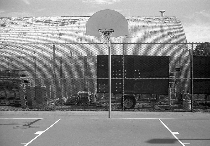 A black and white photograph of a basketball hoop with a road sign with the words San Pablo displayed on it.